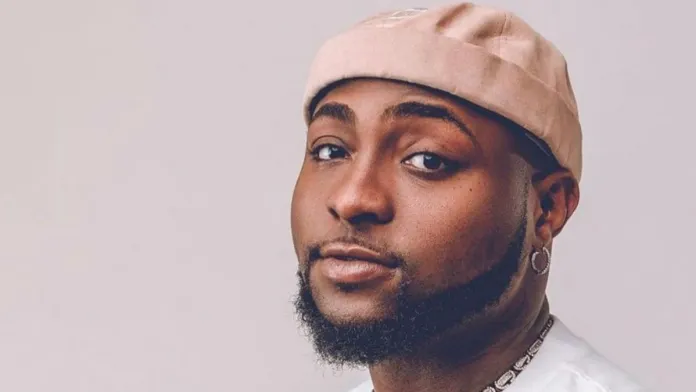 A man with a beard and mustache wears a light pink beanie, a white shirt, and a hoop earring, looking towards the camera with a neutral expression—his style echoing the cool confidence of Afrobeats GOATs like Davido.