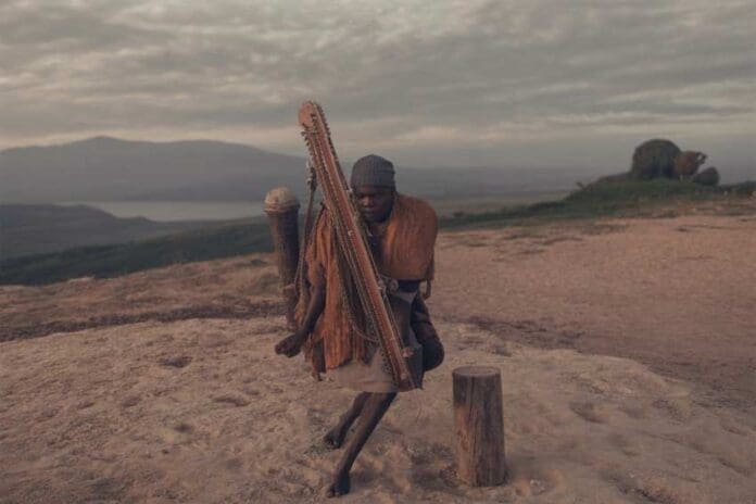 A person stands outdoors on rocky ground in Lagos, holding a large stringed musical instrument, with a cloudy sky and distant hills in the background—capturing the spirit of an African music festival during Detty December Fest.