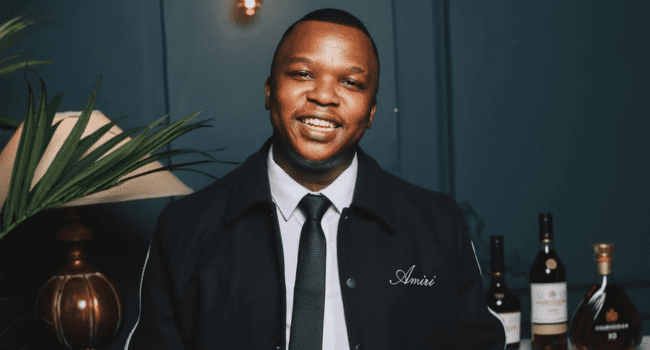 A man in a dark jacket and tie stands indoors, smiling at the camera, as several bottles and a lamp rest on the table beside him—a nod to SA Hip Hop vibes and global heights of style.