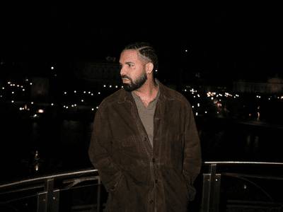 A man with a trimmed beard and short hair stands on a balcony at night in a brown jacket, city lights glowing behind him—perhaps listening to one of Jacaranda FM's most listened-to songs by top artists in SA.