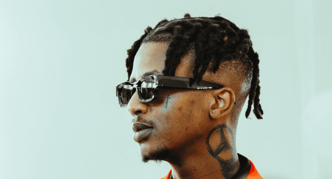 A man with short dreadlocks, wearing black sunglasses and a red shirt, channels Emtee’s bold style with a large peace sign tattoo on his neck and a small face tattoo near his eye.