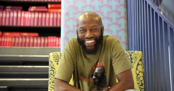 A man with a beard and bald head sits smiling on a patterned chair, in front of a staircase and shelves filled with red books, his expression reflecting the pride of an Eastern Cape local featured in the Daily Dispatch for music awards achievements.