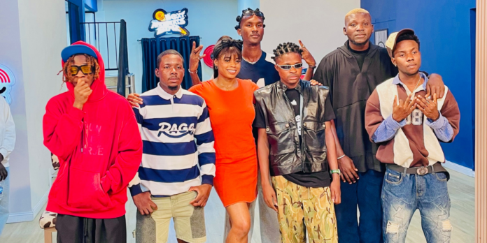 Seven people stand indoors, posing for a group photo; some don streetwear while one in the center wears a bright orange dress, ready to compete in the Next Afrobeats Star contest for a $100,000 prize.