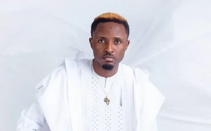 A man wearing a white traditional outfit and necklace stands against a plain white background, looking directly at the camera—Gaise Baba, a prominent Gospel singer on Nigeria’s YouTube music chart.