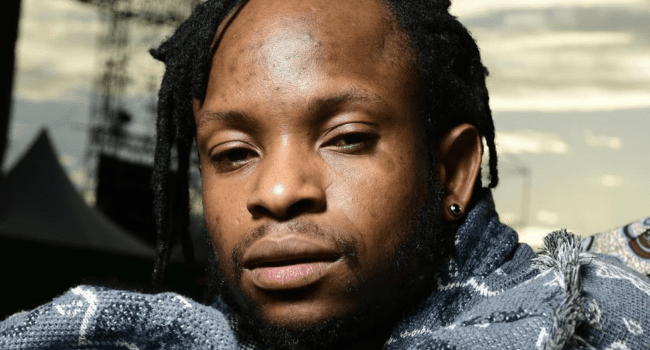 A close-up of a person with short dreadlocks, a beard, and an earring, looking at the camera with a neutral expression. The blurred outdoor background adds depth—reminiscent of cover art for an unreleased joint by Gemini Major.