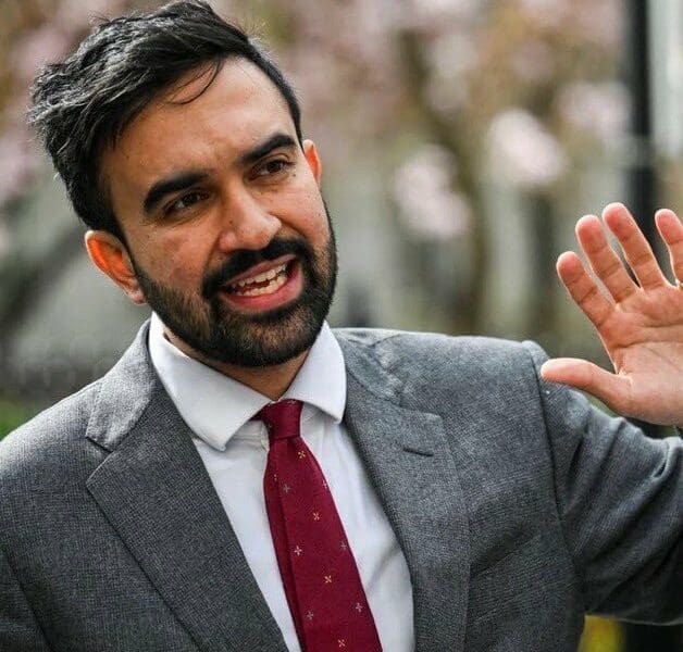 A man in a gray suit and red tie is speaking and gesturing with his right hand outdoors, reminiscent of a New York mayor-elect addressing a crowd.