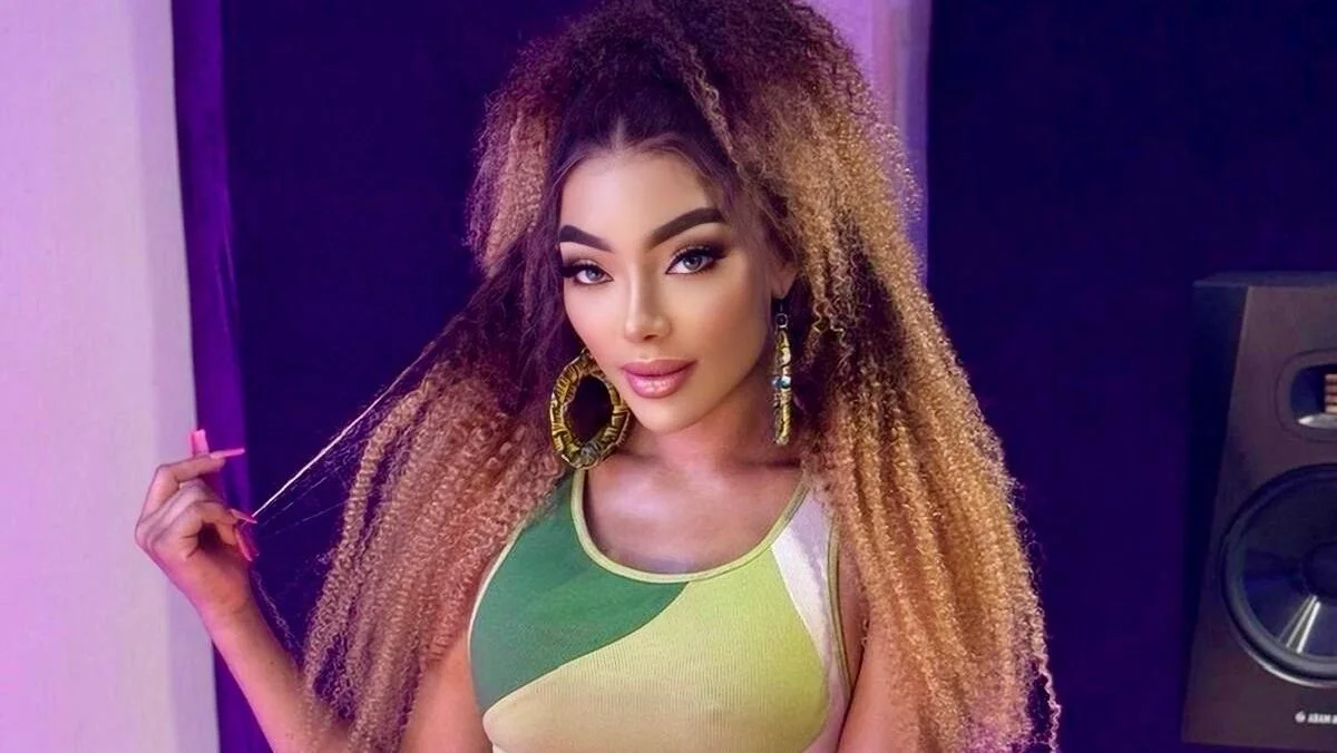 A woman with long curly hair, wearing large hoop earrings and a sleeveless multicolored top, poses indoors in front of a dark background, exuding a vibrant multicultural fusion style.