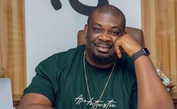 A man with a short beard sits at a desk, resting his head on his hand and smiling. He wears a dark t-shirt, a necklace, and a smartwatch—radiating the confidence of Don Jazzy amid Afrobeats’ global success.