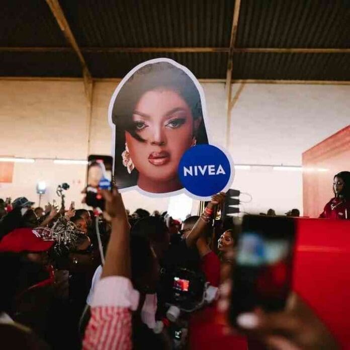 A crowd holds up a large cutout of a woman's face with a Nivea logo during an indoor event featuring DBN Gogo; several people are recording with their phones.