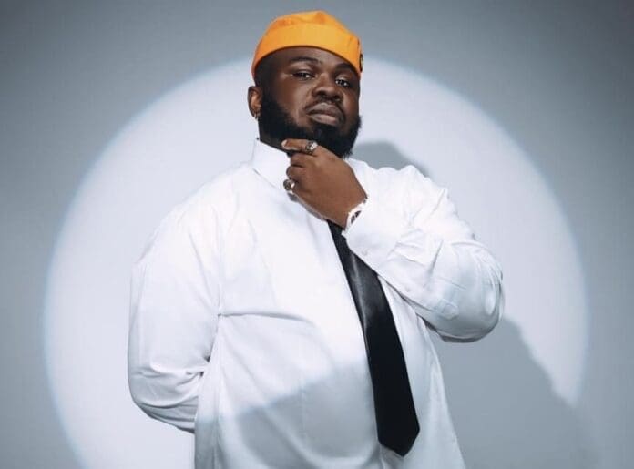 A man wearing a white shirt, black tie, and orange hat stands in front of a light gray background with a circular light pattern, embodying the vibrant energy of contemporary African music.