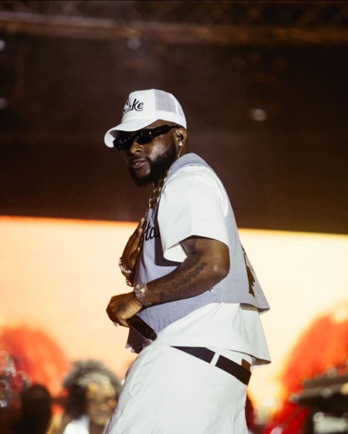 A man wearing sunglasses, a white cap, and light-colored streetwear performs on stage at the 5 Alive Tour in Abuja against a bright, blurred background.