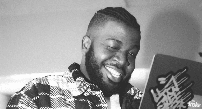 A man with a beard and braided hair smiles and winks at the camera while using a laptop, reminiscent of J Smash channeling positivity even after losing his grandmother.