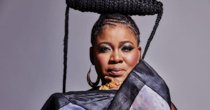 A woman with braided hair styled in a large circular shape on top of her head, wearing dark eye makeup, gold jewelry, and a patterned garment, poses against a plain background, evoking the spirit of Sankofa Festival 2026.
