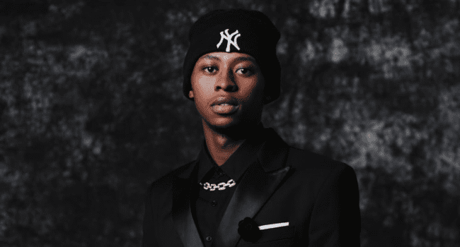 A person wearing a black suit, chain necklace, and a black beanie with a white "NY" logo stands in front of a dark, textured background, channeling strong SA Hip Hop vibes—perfect for K-Rev's Debut Album Release.