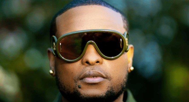 A person with short hair, wearing large green sunglasses and gold earrings, faces the camera outdoors with a blurred background—capturing the bold style of a K.O concert, perfect for Young Fans at Live At The Hill.