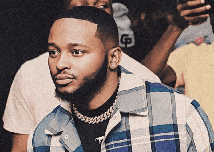 A man with a trimmed beard, wearing a blue plaid shirt and a silver chain necklace, stands in front of a blurred crowd at Mdzimba View—capturing the vibrant spirit often featured in Eswatini Positive News.