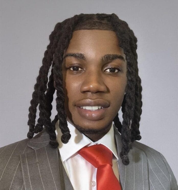 A man in a pinstriped gray suit, white shirt, and red tie poses for a portrait against a plain gray background. With shoulder-length twisted hair, he exudes the cool confidence often seen in Soul Music and Afro Pop artists.