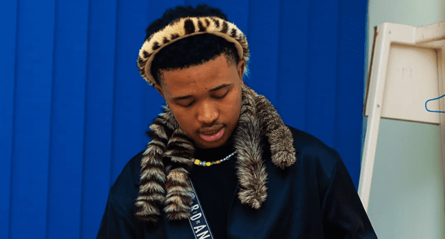 A person wearing a traditional animal print headband and a fur scarf stands in front of a blue vertical blind background, looking down—capturing the moody style of Lowfeye's NOSANGE debut album.
