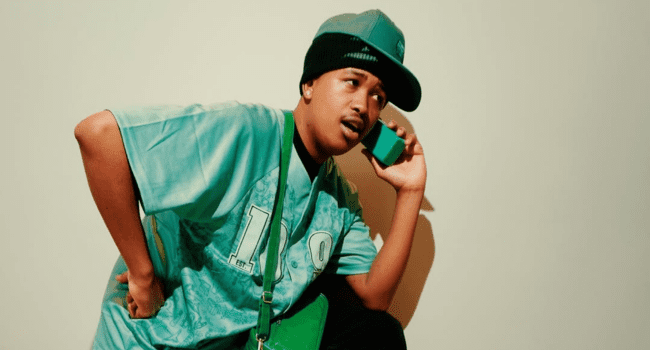 Person wearing a teal shirt and cap, holding a green object to their ear as if talking on a phone, striking a pose inspired by the Umfana KaMakhulu album, standing against a plain background.