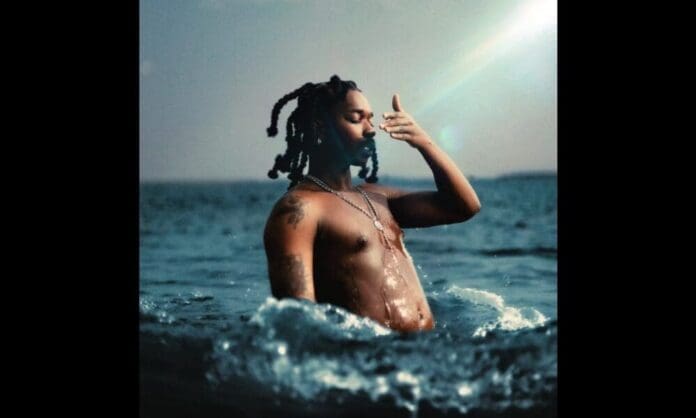 A shirtless person with tattoos stands waist-deep in water, touching their face, with sunlight and waves around them—capturing a vibe reminiscent of BellaNaija’s striking coverage of Nigerian music albums set for 2025.