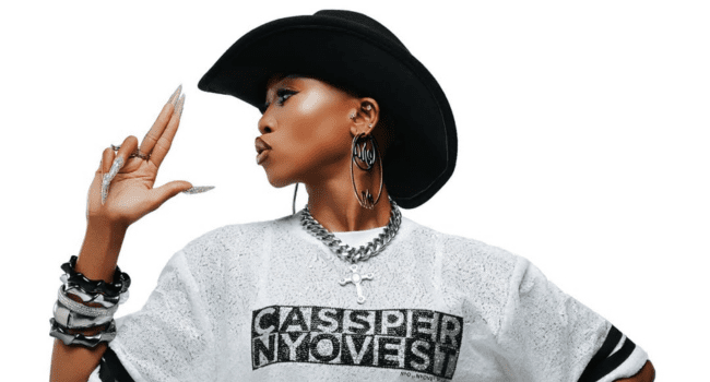 A person wearing a black cowboy hat, large hoop earrings, and a “Cassper Nyovest” t-shirt poses with fingers pointed in a gun gesture—celebrating the Fill Up Toyota Stadium vibe.