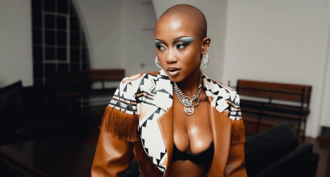 A woman with a shaved head wears bold makeup, a patterned jacket with brown fringe, a black bra, hoop earrings, and layered necklaces, channeling Moozlie's signature style at Cotton Fest while standing indoors.