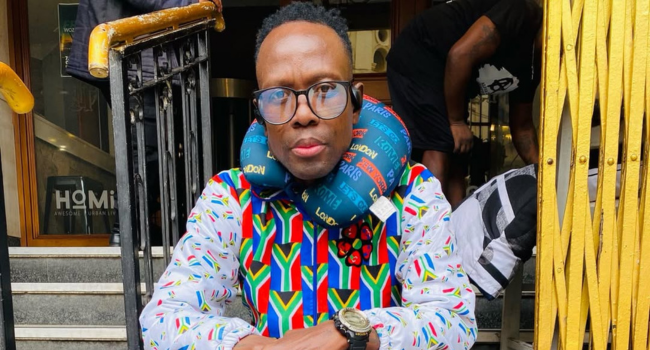 Rashid Kay, wearing glasses and a colorful South African flag-patterned shirt, sits on stairs with a travel neck pillow around his neck—reflecting the style recognized at the Timeline Radio Awards for Honorary Glory.