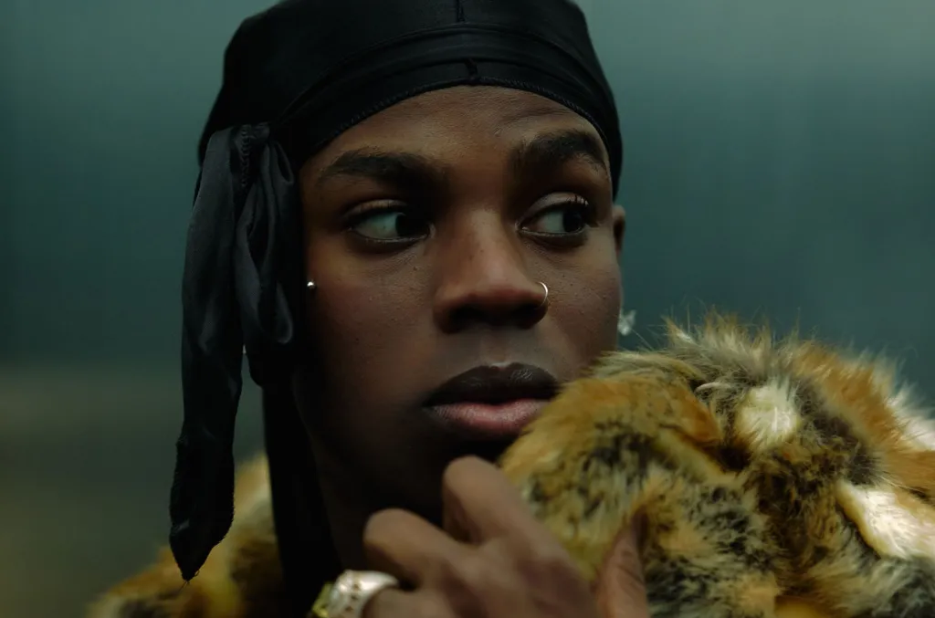 A person wearing a black durag and a nose ring looks to the side, holding a fur-collared garment close to their face, embodying the bold style seen in African Fresh Picks on Billboard.