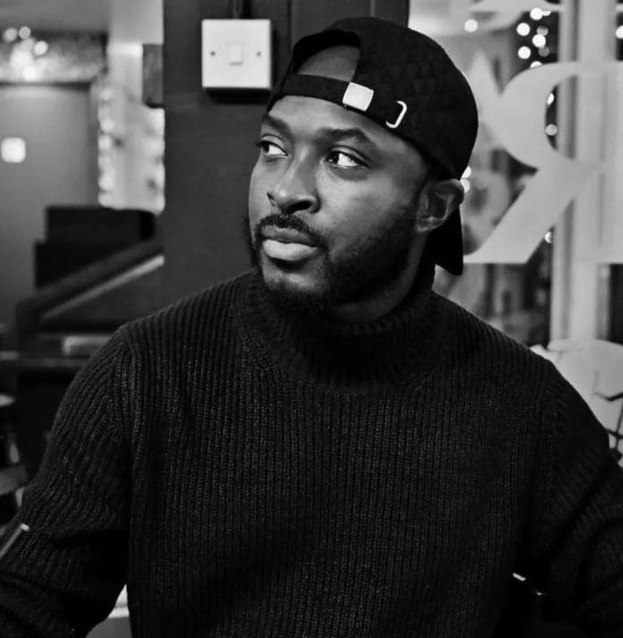 A man in a dark sweater and backward cap sits indoors, looking to the side. The black and white photo captures a contemplative vibe, reminiscent of Young Rob's Afrobeats style.