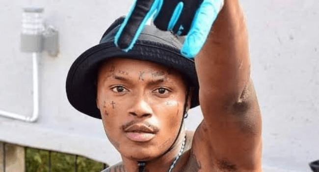 A person wearing a black bucket hat and blue glove raises their arm, showing tattoos on their face and arm, standing outdoors near a wall—reminiscent of Shebeshxt amid his recent legal woes.