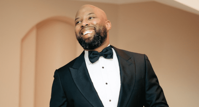 A man in a black tuxedo and bow tie smiles while standing indoors in front of a light-colored wall, reminiscent of DJ Warras at an exclusive event.