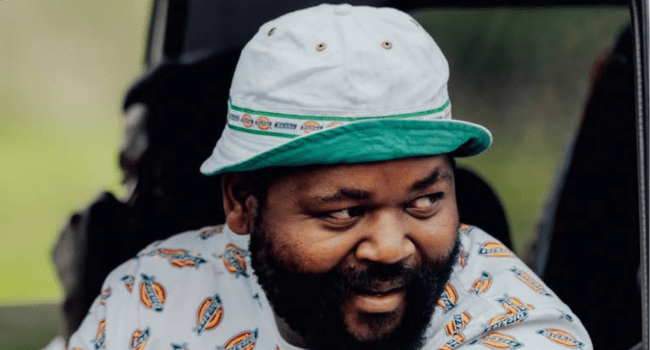 A man with a beard wearing a white hat with a green trim and a patterned shirt looks to the side, smiling—reminiscent of Sjava from the INkanyezi NeZinkanyezi album.