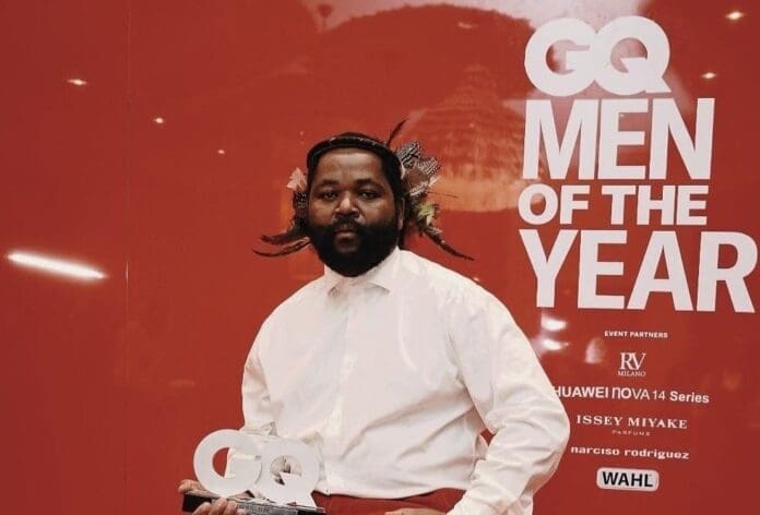 A man in a white shirt and red pants holds a GQ award in front of a red backdrop with the text "GQ Men of the Year," celebrating SJAVA as the Music Excellence Winner, featured by SA Music News Magazine.