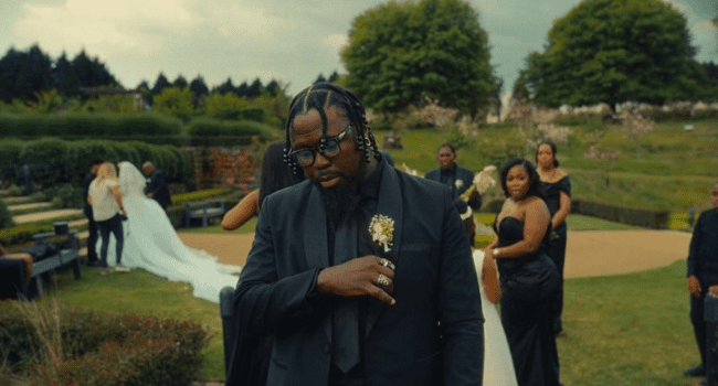 A man in a dark suit with sunglasses stands outdoors at a formal marriage event, with other guests and a bride in the background—exuding style reminiscent of Stilo Magolide.