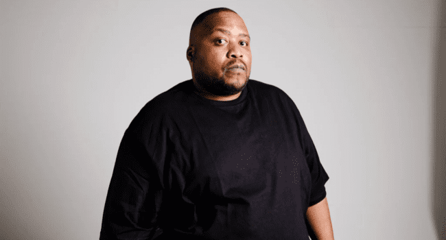 A man with short hair and a beard stands against a plain background, wearing a black t-shirt and looking toward the camera with a neutral expression, reminiscent of musician Stogie T from the Four Horsemen collective.