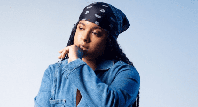 A person with long braids, channeling Tasha Hendrixx’s signature style, wears a black patterned bandana and blue denim shirt while posing against a plain light background.