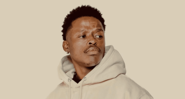 A man wearing a cream-colored hoodie looks to the side against a plain beige background, reflecting on career support and inspiration from icons like Riky Rick.