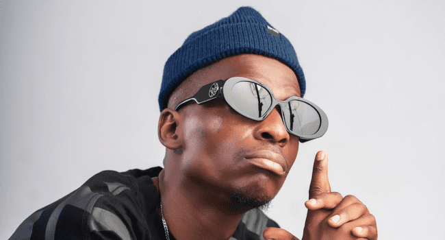 A person wearing a blue beanie, dark sunglasses, and a black shirt holds up one finger while looking slightly upward, channeling the vibe of a Spotify "200 Bars" session.