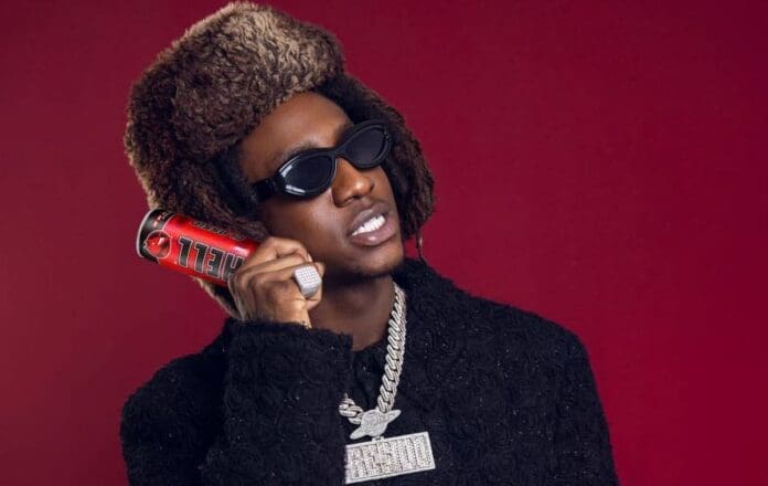 A man wearing a furry hat, dark sunglasses, and heavy jewelry holds a red energy drink can to his ear against a red background, channeling the bold style of Shallipopi, the 2025 TikTok Awards Artist of the Year.