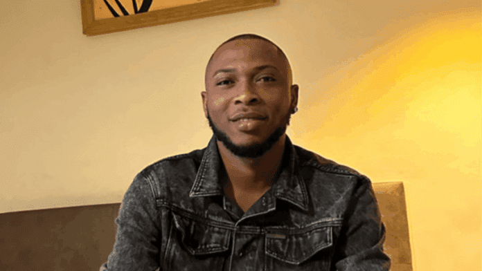 A man wearing a dark denim jacket sits indoors on a couch, facing the camera, with a warm light and framed art behind him—perhaps reflecting on Eliana's new single from KingSai.