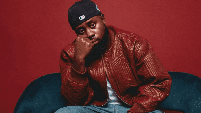 A man wearing a red leather jacket, blue jeans, and a black cap sits on a blue couch against a red background, resting his face on his hand—channelling the cool style of KJ Spio.