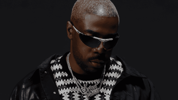 A man with bleached blond hair wearing dark sunglasses, a black and white patterned sweater, a black jacket, and silver jewelry poses against a dark background—channeling the bold style of James BKS’s Deluxe EP “See Us Rise.”.