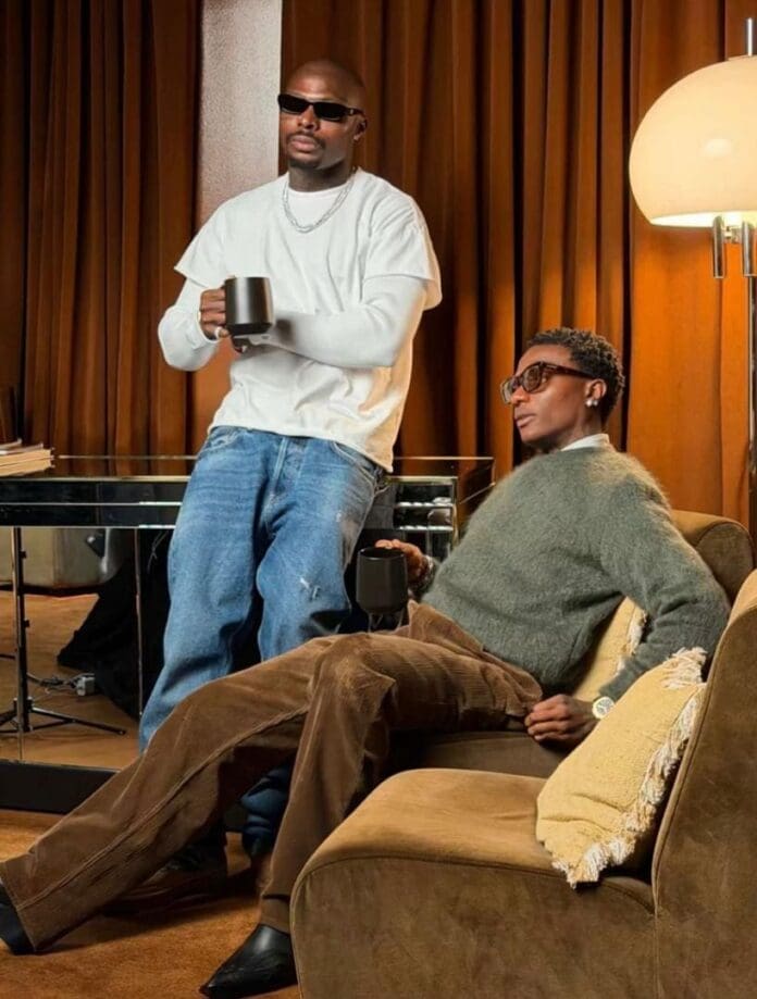 Two men indoors, one standing and one sitting on a sofa, both holding black mugs. The room has brown curtains, a glass table, and a floor lamp, with TVC News playing softly in the background.