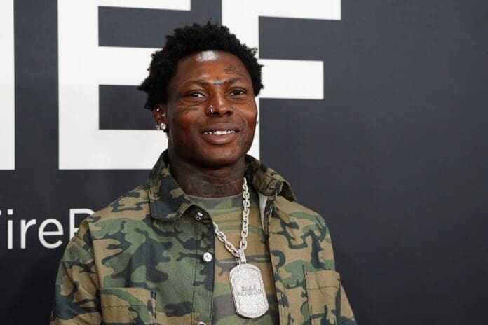 A man in a camouflage jacket and chain necklace stands in front of a black and white background, smiling at the camera, embodying the vibrant spirit of Afrobeats culture.
