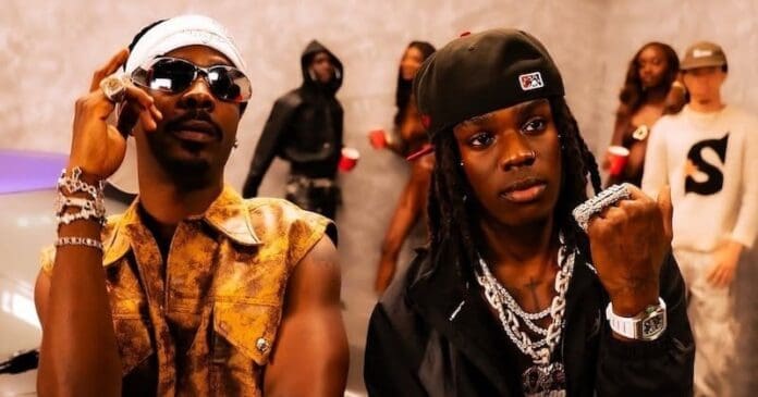 Two men in stylish outfits and jewelry stand in the foreground at a party, channeling the vibrant energy of Rema, with several people and a car visible in the background.