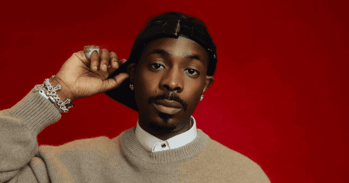 A man in a beige sweater and white collared shirt poses against a red background, wearing a backward black cap and silver jewelry, channeling the vibrant energy of Young Jonn's Blue Disco album.