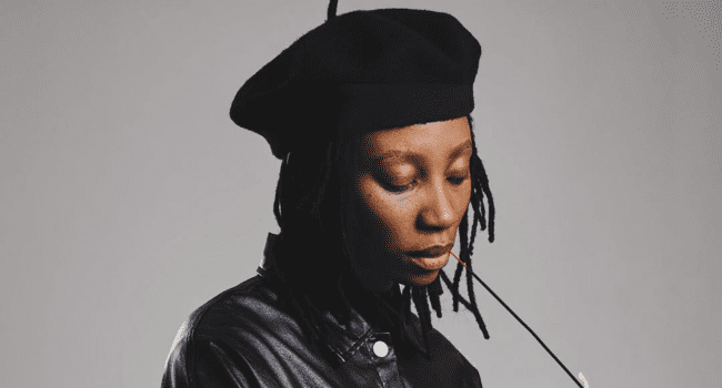 A person wearing a black beret and black leather jacket looks down while holding a pair of glasses near their mouth, embodying the bold spirit of SA Hip Hop.