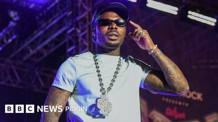 A Nigerian singer wearing sunglasses, a cap, and a large chain necklace performs on stage with colorful lights at the Asake concert Kenya. The BBC News Pidgin logo is visible in the corner.