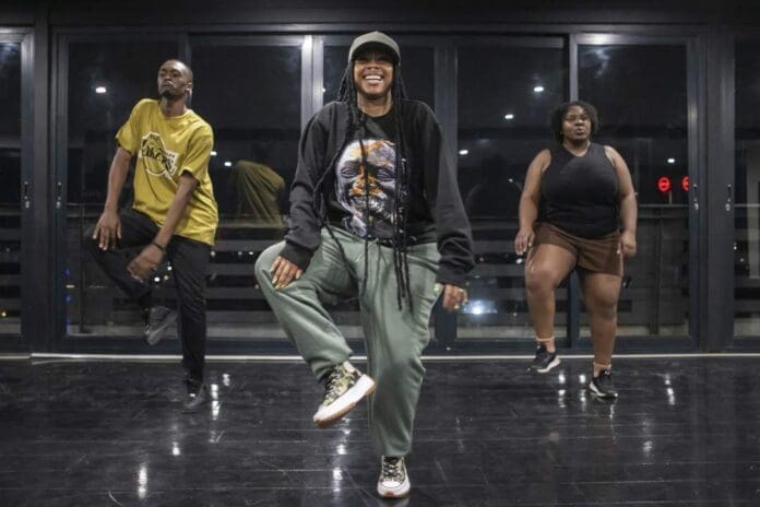 Three people stand on a shiny studio floor, lifting one knee as they practice an amapiano dance routine in front of large windows at night.
