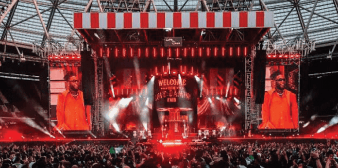 A Nigerian Afrobeats performer in an orange outfit sings on a large, brightly lit stage with red lighting, video screens, and a crowd watching inside a stadium.